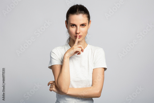 Young beautiful girl making silence gesture, asking not tell anyone secret, on light grey studio background
