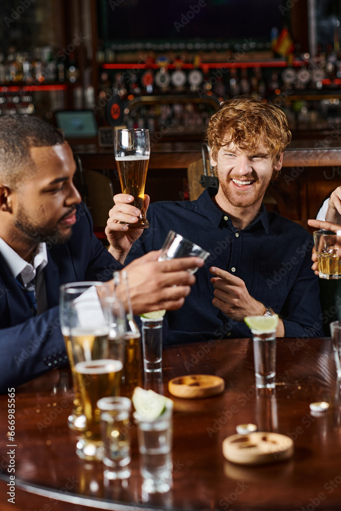 cheerful redhead man pointing at friend while having drinks after work in bar, interracial friends