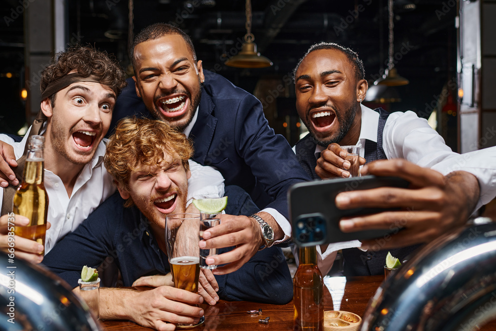 excited multiethnic colleagues in formal wear taking selfie on smartphone in bar after work