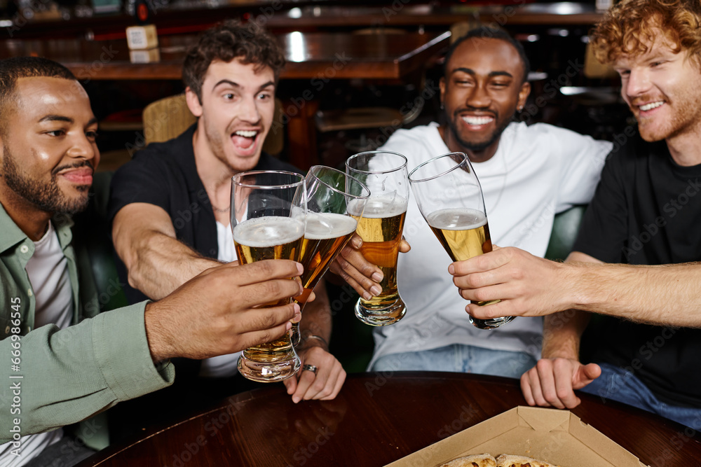 four excited multicultural friends clinking with glasses of beer in bar, men on bachelor party