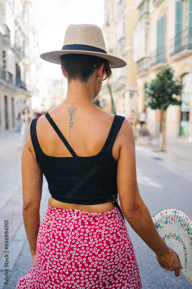 Back of tattooed woman with hat and hand fan walking down an Andalusian street.