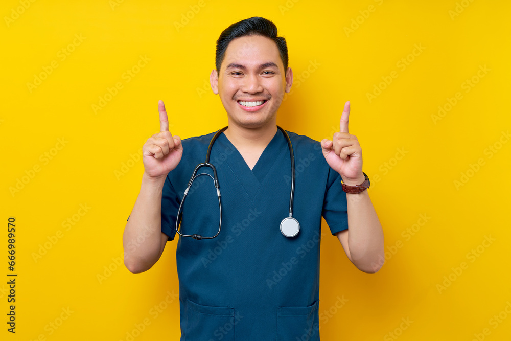 Smiling professional young Asian male doctor or nurse wearing a blue uniform and stethoscope pointing finger up on copy space isolated on yellow background. Healthcare medicine concept Smiling professional young Asian male doctor or nurse wearing a blue uniform and stethoscope pointing finger up on copy space isolated on yellow background. Healthcare medicine concept