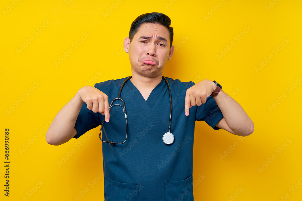 Unhappy professional young Asian male doctor or nurse wearing a blue uniform and stethoscope pointing finger down on copy space isolated on yellow background. Healthcare medicine concept Unhappy professional young Asian male doctor or nurse wearing a blue uniform and stethoscope pointing finger down on copy space isolated on yellow background. Healthcare medicine concept