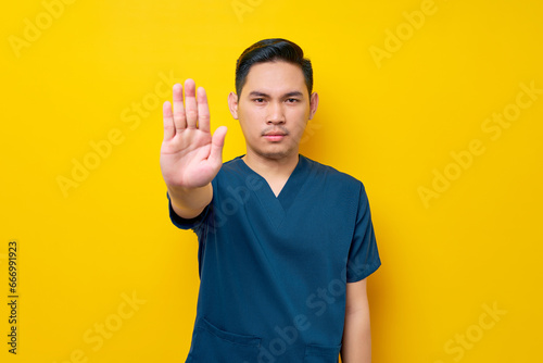 Serious professional young Asian male doctor or nurse wearing a blue uniform makes a stop gesture with his hands isolated on yellow background. Healthcare medicine concept