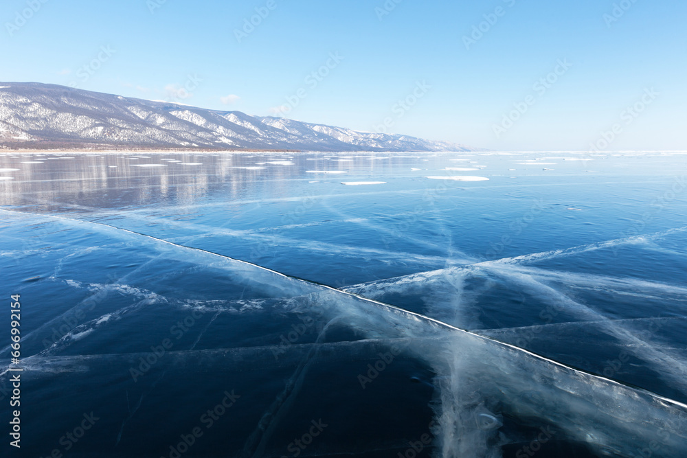 Fototapeta premium Scenic unusual winter landscape of frozen Baikal Lake on sunny February day. Small Sea is covered with beautiful transparent blue ice with cracks. Endless icy desert to horizon. Winter travel 