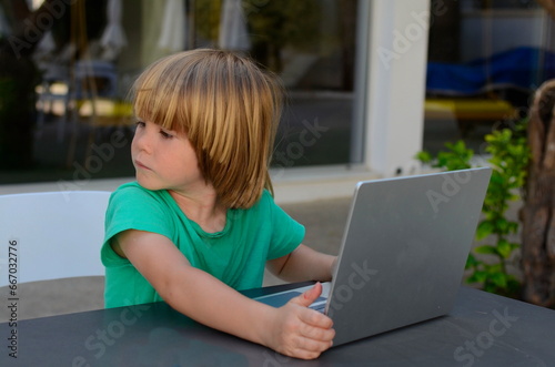 A little boy sits at a laptop, doing his homework. Preschooler studying, boy busy with computer outside in cafe