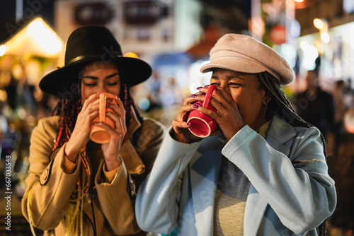 African mother and daughter having drinking hot chocolate outdoor at winter market - Focus on senior female hand holding cup