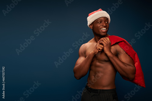 smiling african american man with muscular torso and christmas bag looking away on dark blue © LIGHTFIELD STUDIOS