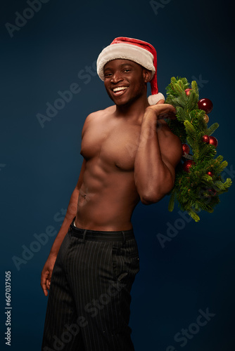 shirtless sportive african american model in santa cap posing with christmas wreath on dark blue