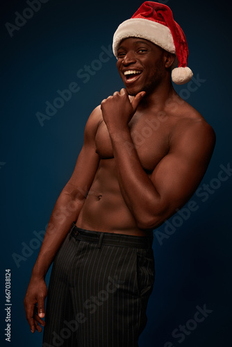 muscular and shirtless african american man in santa hat laughing at camera on navy blue backdrop