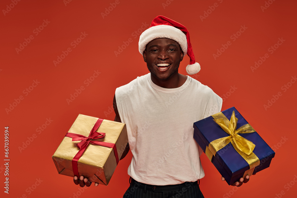 joyful african american man in christmas hat and white tank top posing with festive presents on red joyful african american man in christmas hat and white tank top posing with festive presents on red