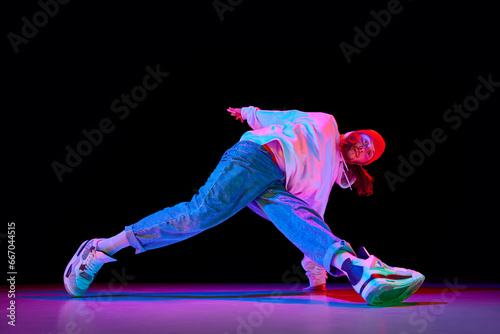 Young man in sportswear in motion, dancing breakdance isolated over black studio background in neon light. Concept of contemporary dance, street style, fashion, hobby, youth. Ad