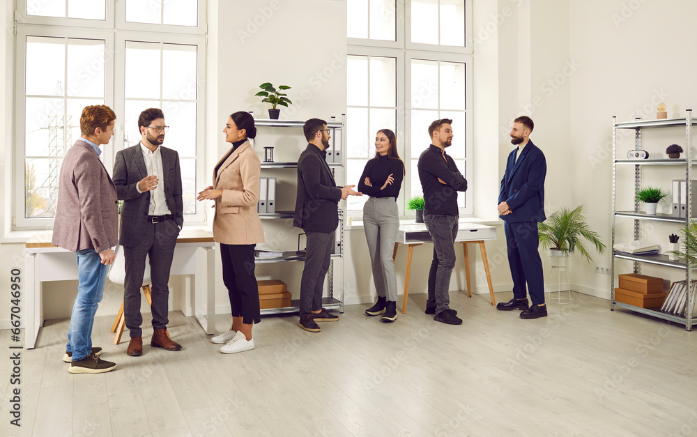Young people communicating at a work meeting in the office. Group of men and women standing in a modern office interior, talking, discussing work and sharing business ideas. Communication concept - obrazy, fototapety, plakaty 