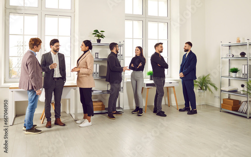 Young people communicating at a work meeting in the office. Group of men and women standing in a modern office interior, talking, discussing work and sharing business ideas. Communication concept