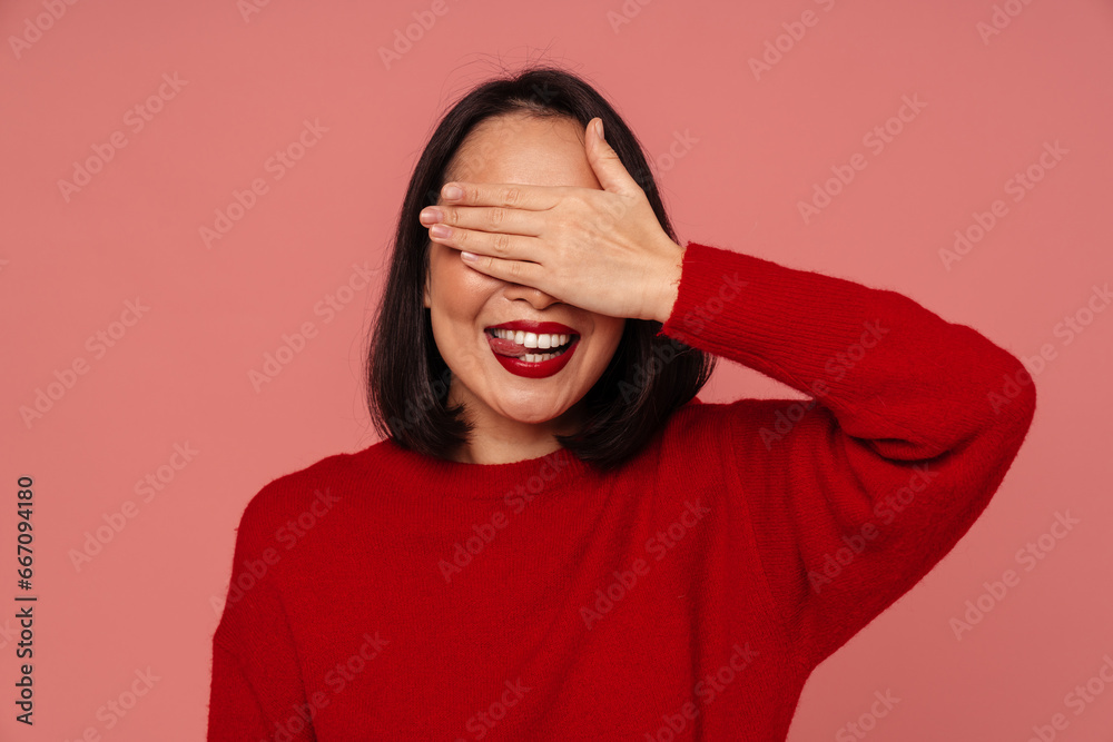 Cheerful woman covering her eyes and showing tongue while standing isolated over pink wall Cheerful woman covering her eyes and showing tongue while standing isolated over pink wall