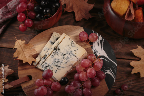Cheese platter served with autumn fruits and dry leaves on dark wooden background. Appetizer of blue cheese on cutting board with grapes. Top view