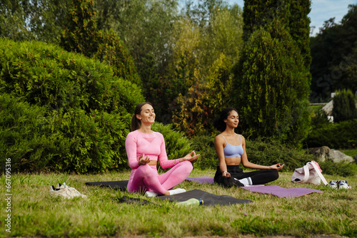 Two young women sitting on yoga mats in lotus pose and meditating in green park
