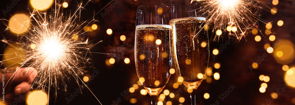 Toasting champagne for festive celebrations. Glasses of sparkling wine in front of golden bokeh lights and glowing sparklers. Horizontal background for christmas, new year, parties and festive events. - obrazy, fototapety, plakaty 