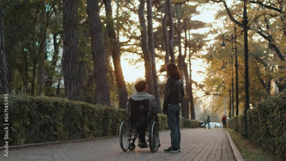Disabled man in a wheelchair and a woman stand on a path in an autumn city park and talk during a walk together, view from behind. Concept of acceptance, love and support for people with disabilities Disabled man in a wheelchair and a woman stand on a path in an autumn city park and talk during a walk together, view from behind. Concept of acceptance, love and support for people with disabilities