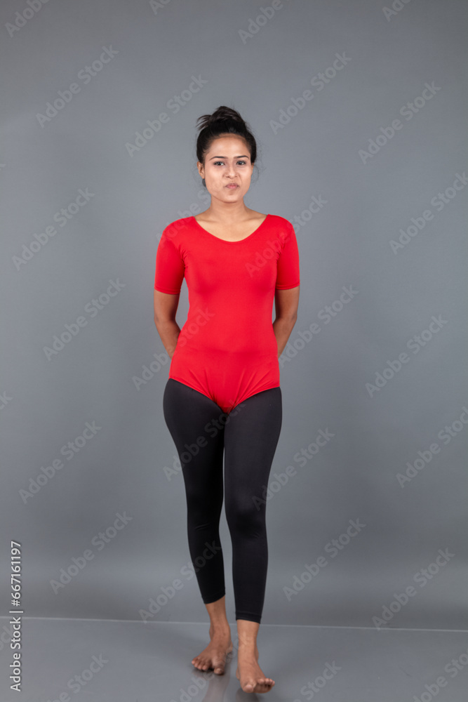 Healthy young woman in red t-shirt and black leggings doing yoga and stretching exercises isolated on grey background
