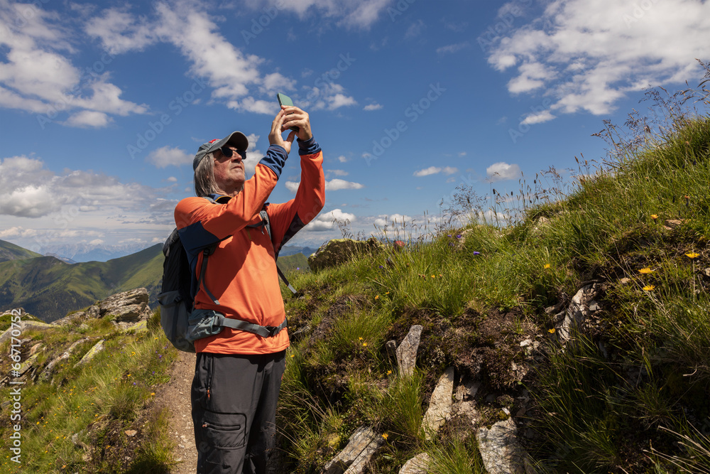 Gray-haired man in cap and backpack taking pictures of mountain landscape against bright sky, Austria