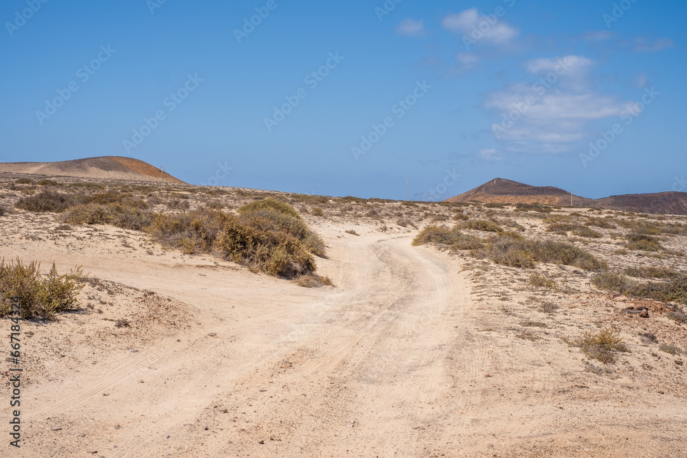 Desert landscape of white sand and desert shrubs. Dirt road and mountains in the background. Sky with big white clouds. Lanzarote, Canary Islands, Spain.