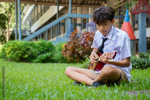 Asian schoolboy in school uniform playing ukulele or acoustic guitar lonely in school park, asian boy with popular intruments concept. photo