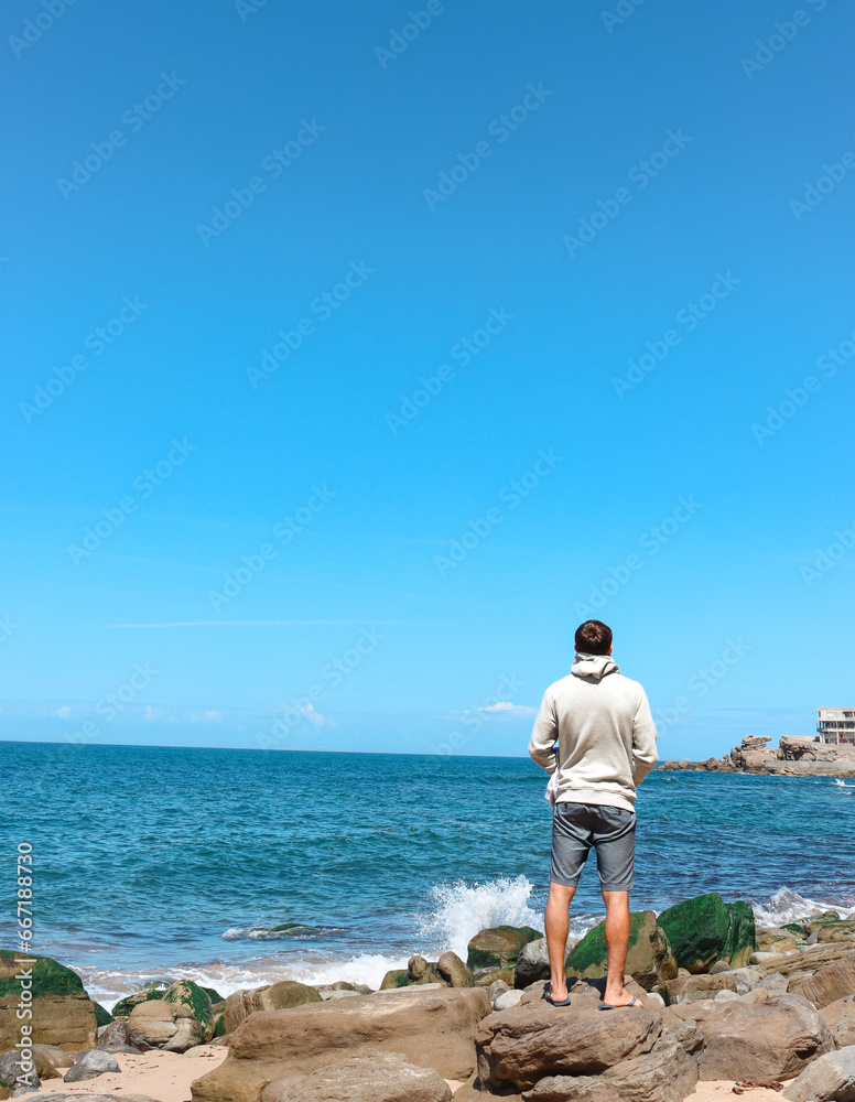 Young man traveler contemplating in a peaceful place in fornt of the ocean viewed from the back. Portugal Solo traveler.
