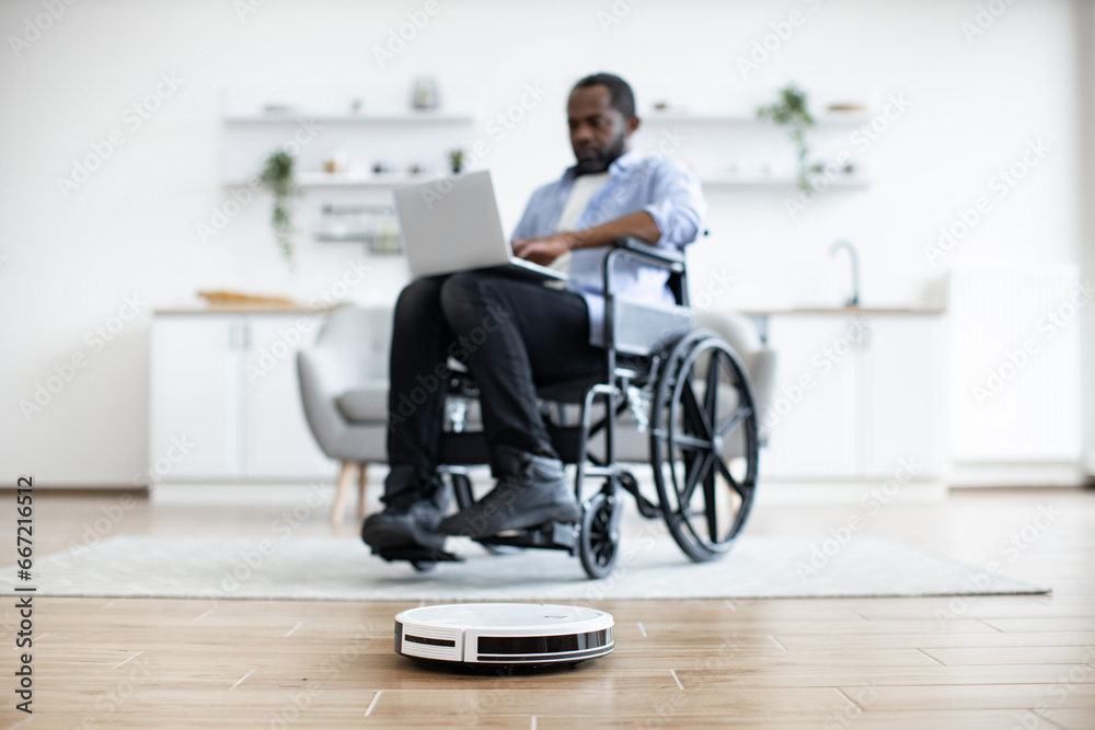 Selective focus of robotic vacuum removing dust from carpet while african homeowner with mobility impairment using laptop. Freelancer in wheelchair leading comfortable lifestyle by using tech gadgets Selective focus of robotic vacuum removing dust from carpet while african homeowner with mobility impairment using laptop. Freelancer in wheelchair leading comfortable lifestyle by using tech gadgets