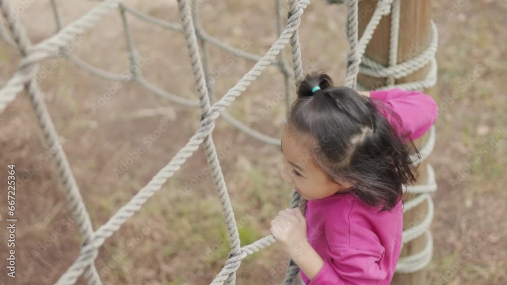 A curious and adorable Asian girl, her face with a cheerful smile, is seen climbing a rope net with carefree enthusiasm. It's training outdoor, exercising, or enjoying the leisure of kid and child. A curious and adorable Asian girl, her face with a cheerful smile, is seen climbing a rope net with carefree enthusiasm. It's training outdoor, exercising, or enjoying the leisure of kid and child.