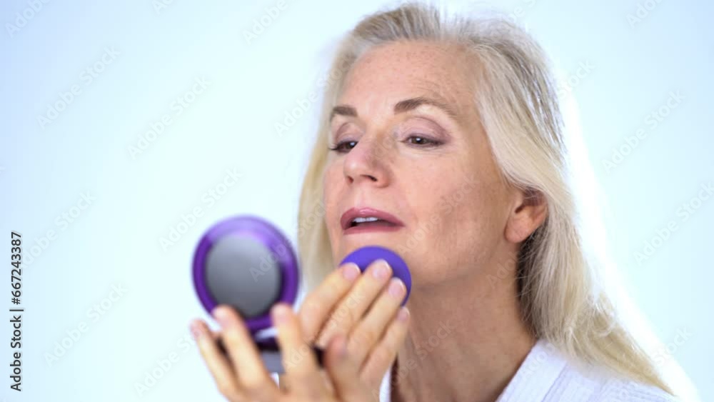 Closeup portrait of 50s, 60s, mature middle aged woman looking in mirror applying natural makeup powder foundation with cosmetic sponge on clean face skin smoothing wrinkles, on white background. Closeup portrait of 50s, 60s, mature middle aged woman looking in mirror applying natural makeup powder foundation with cosmetic sponge on clean face skin smoothing wrinkles, on white background.