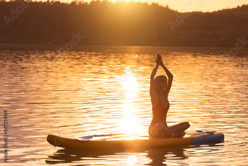 Young woman in yoga pose practicing on paddle sup surfboard during sunrise or sunset. © zinkevych