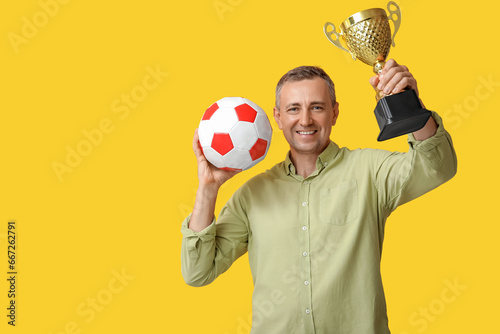 Handsome mature man with gold cup and soccer ball on yellow background © Pixel-Shot