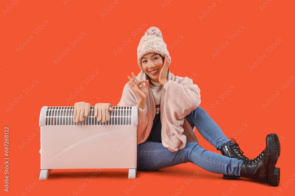 Young Asian woman in winter clothes with radiator sitting on orange background
