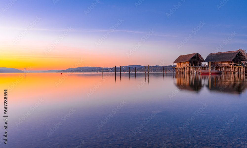 A serene sunrise scene by a tranquil lake with a small house on the shore. The sky is a beautiful gradient of warm colors reflected in the calm water, creating a peaceful atmosphere.