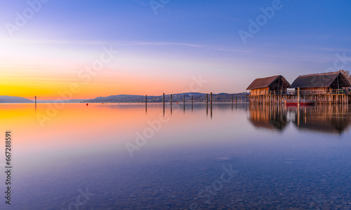 A serene sunrise scene by a tranquil lake with a small house on the shore. The sky is a beautiful gradient of warm colors reflected in the calm water, creating a peaceful atmosphere. © bersch28