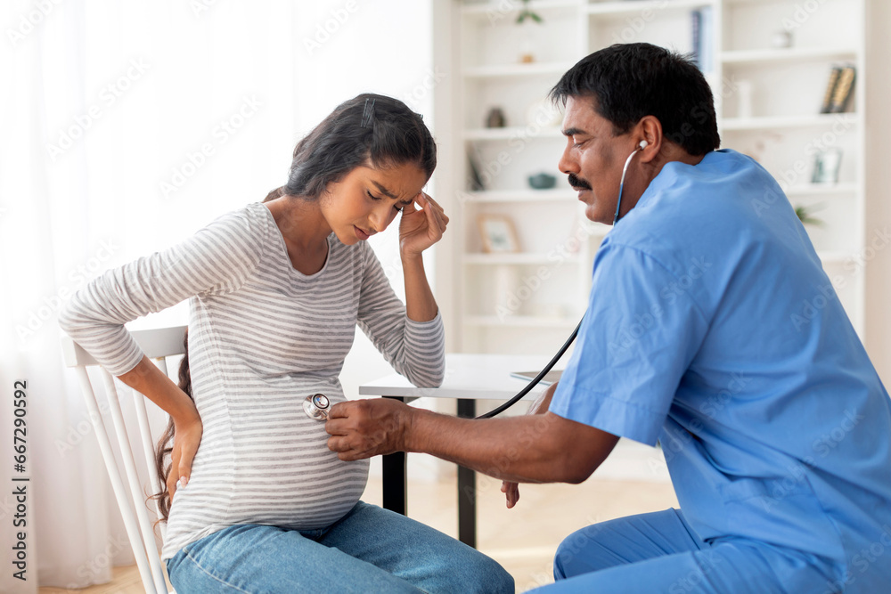 Obraz premium Indian Male Gynecologist With Stethoscope Examining Belly Of Sick Pregnant Female Patient