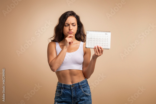 Confused millennial woman with calendar on beige background