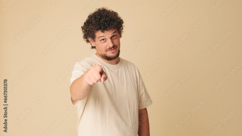 Smiling guy with curly hair dressed in beige t-shirt, shows finger forward looking at camera on beige background in the studio Smiling guy with curly hair dressed in beige t-shirt, shows finger forward looking at camera on beige background in the studio