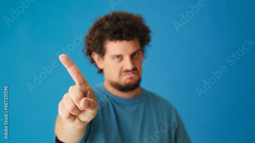 Close-up of young man's hand showing no gesture, waving finger showing negative answer, not allowed, refusal gesture isolated on blue background in studio, background blurred © Andrii Nekrasov