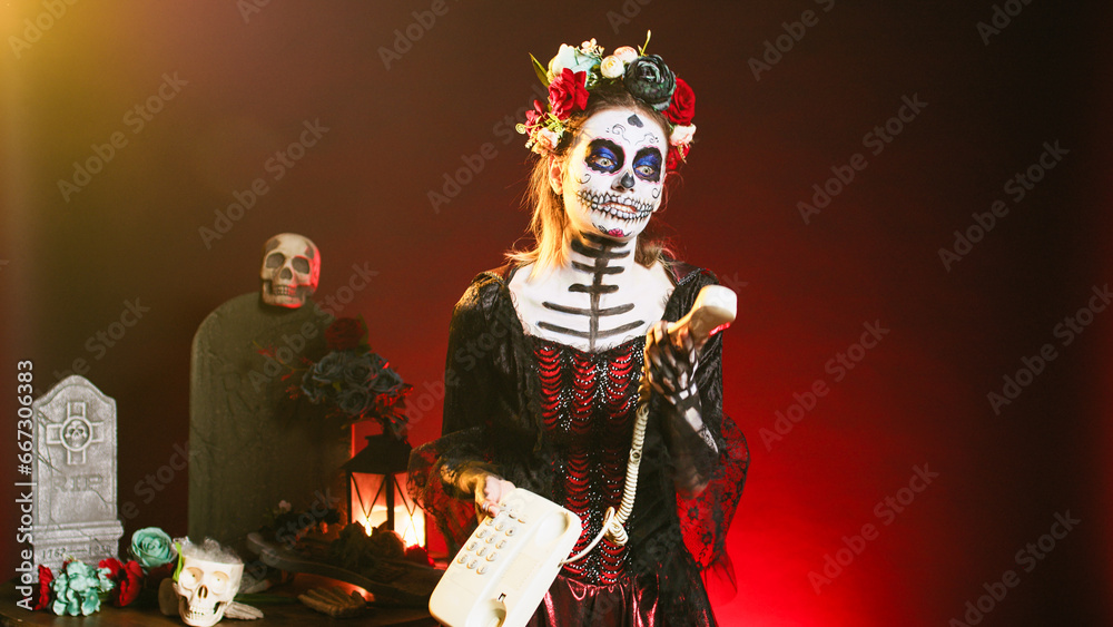 Spooky woman answering landline phone with cord, talking on office telephone while she has skull make up and body art. Portraying santa muerte lady of death on dios de los muertos celebration.