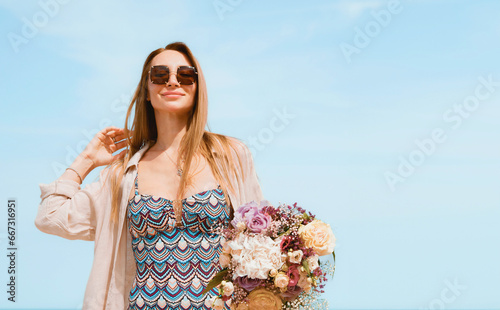 Joyful Young Woman Holding a Bouquet of Flowers on a Beach with Blue Sky Background