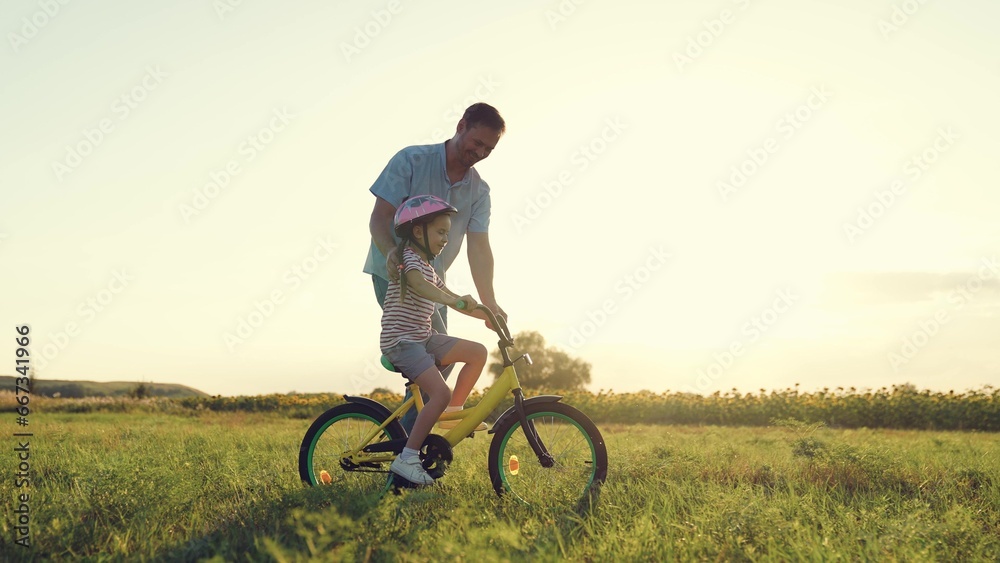 Smiling father teaches preschooler daughter to ride bicycle in summer field