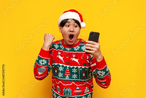 young asian man in christmas sweater and santa hat uses smartphone and celebrates victory photo