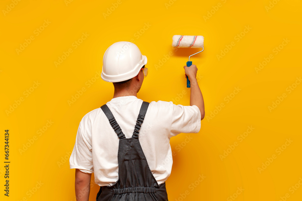 painter man in uniform works and paints the wall yellow with roller, repairman in hard hat and overalls - obrazy, fototapety, plakaty 