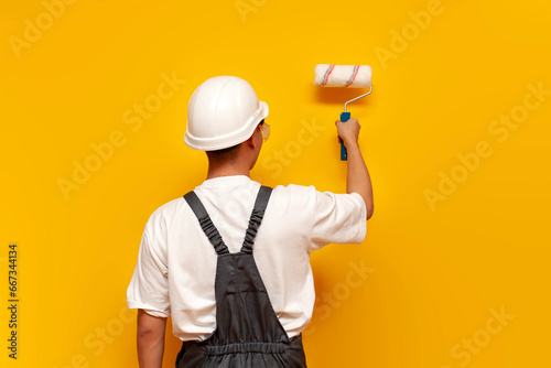 painter man in uniform works and paints the wall yellow with roller, repairman in hard hat and overalls photo