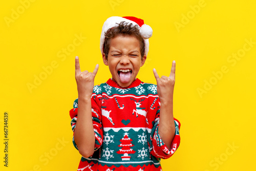 crazy african american boy in christmas sweater and santa hat shows tongue and rock gesture on yellow background photo