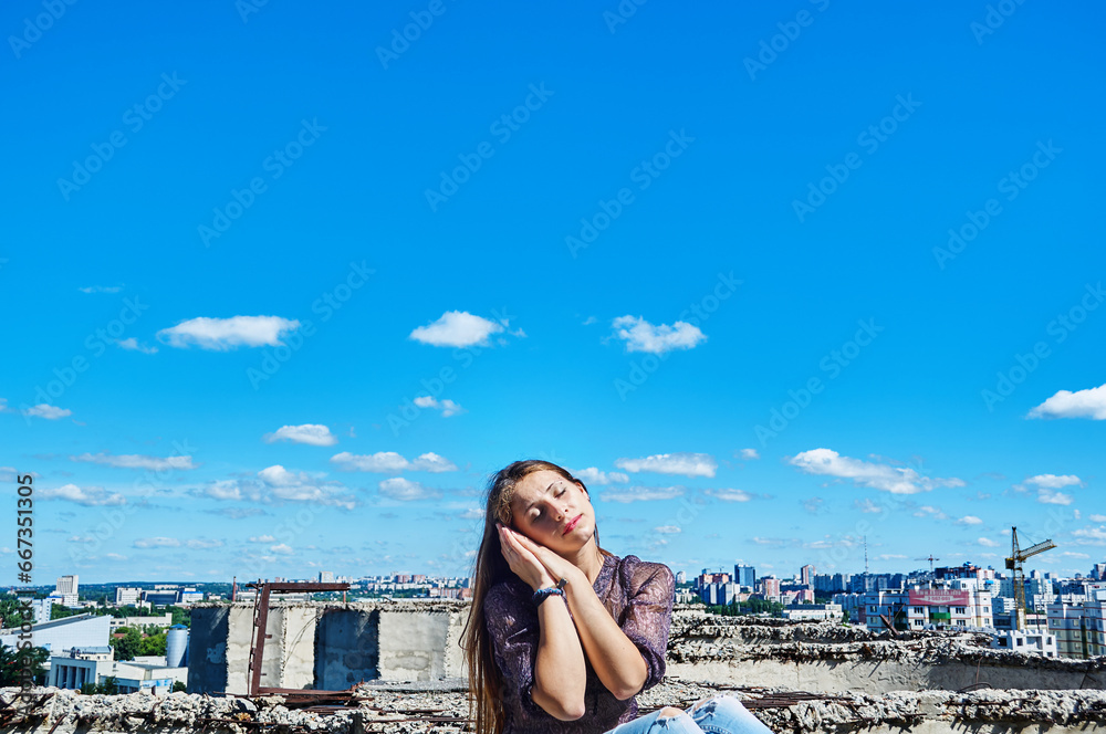 A girl who is not afraid of heights, with a calm face, sits next to a precipice against the backdrop of the city and the blue cloudy sky.