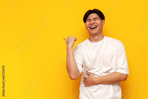 young asian guy in white t-shirt laughing and pointing to the side on yellow isolated background photo