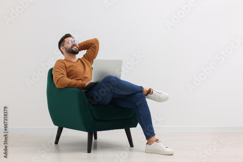 Handsome man with laptop relaxing in armchair near white wall indoors, space for text